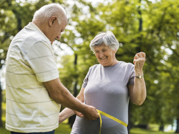 Senior man measuring his wife&#039;s waist with tape measure in the park. Focus is on woman.