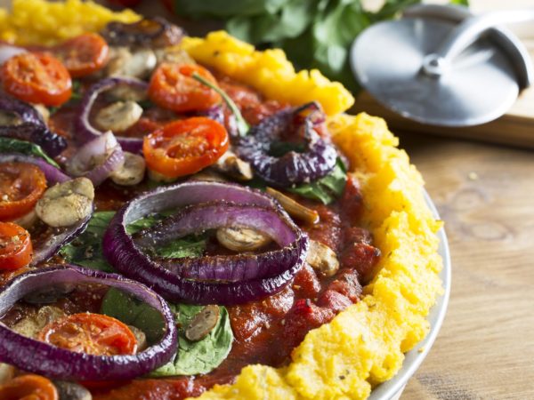 Whole vegan polenta pizza with cherry tomatoes, mushrooms, spinach and red onions.  Close up with pizza slicer out of focus in the background, horizontal orientation.