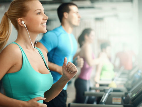 Group of late 20&#039;s people doing treadmill exercise in a local gym. There are three girls and a guy. The closest woman is in sharp focus. She&#039;s listening to mp3 and smiling while doing her workout. She has long blond hair pulled back in a pony tail and wearing green sleeveless tank top. Toned image, side view.