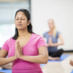 A multi-ethnic group of adults are taking a yoga class together in the gym. They are sitting together and are meditating with their eyes closed on their yoga mats.