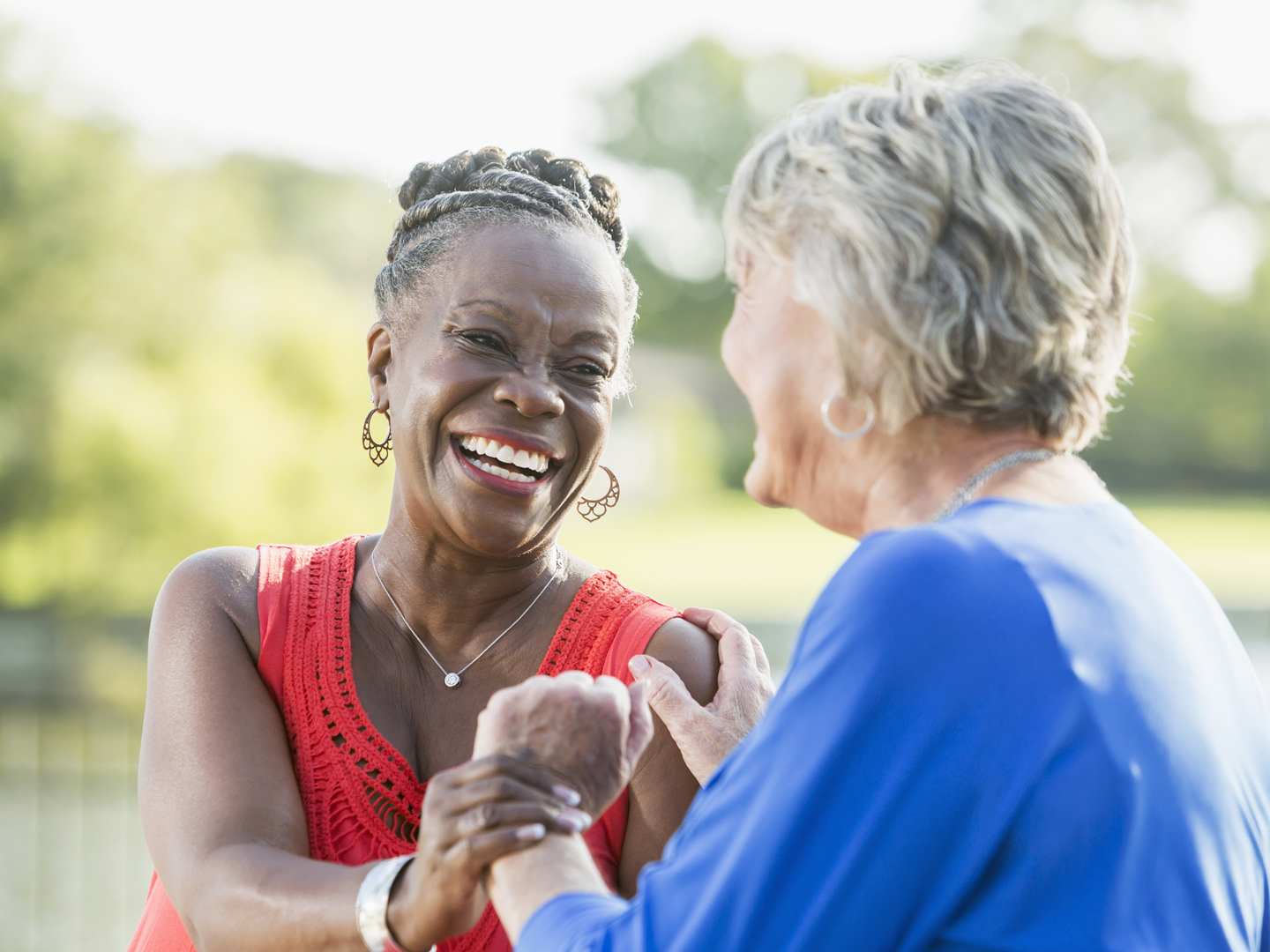 Two senior multi-ethnic woman, one Caucasian and the other African American, sitting in back yard by water enjoying each other&#039;s company. They are looking at each other, laughing.