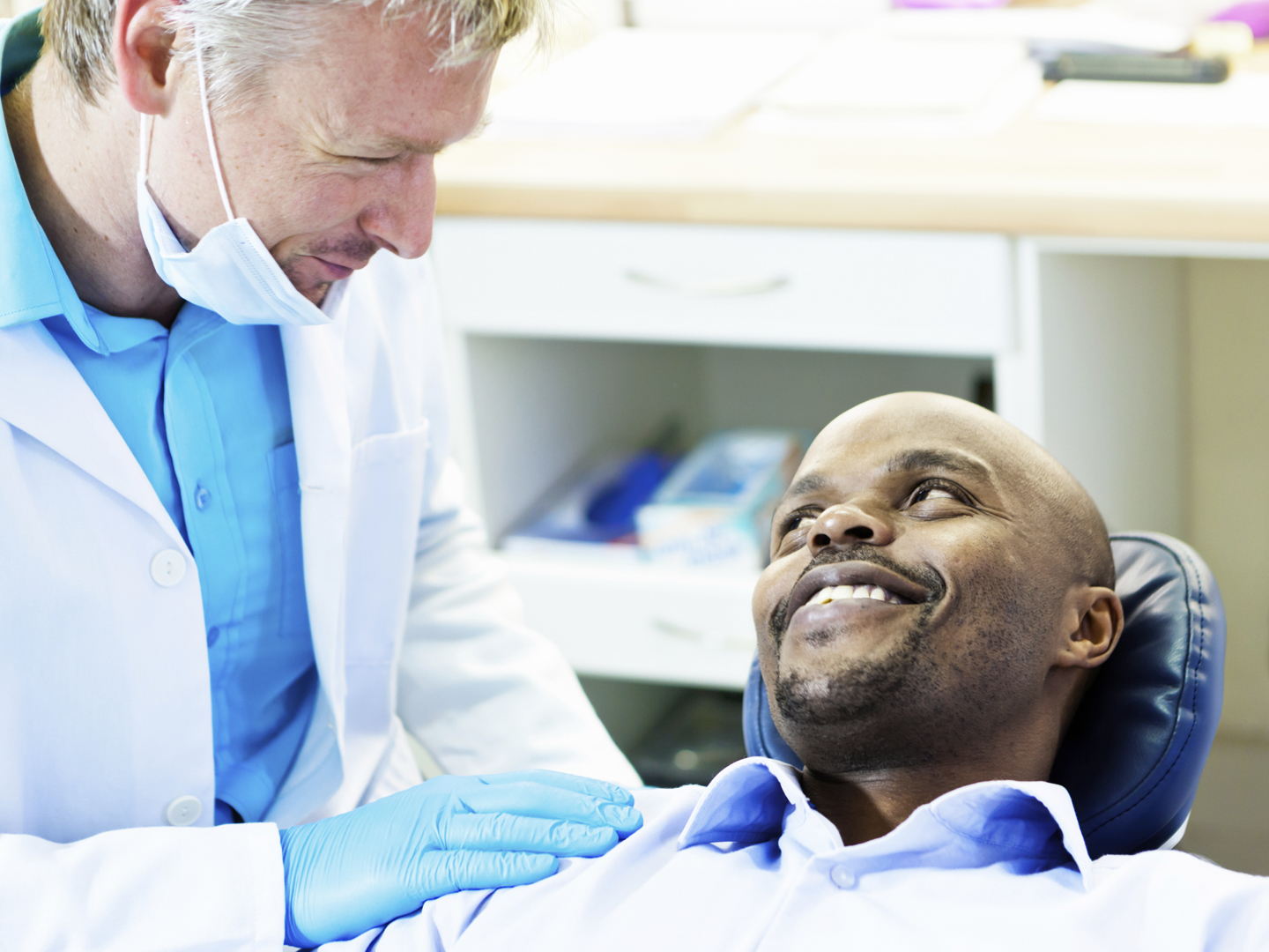 A male patient reclining in the dentist&#039;s chair smiles happily up as his dentist looks down at him, patting his shoulder reassuringly.