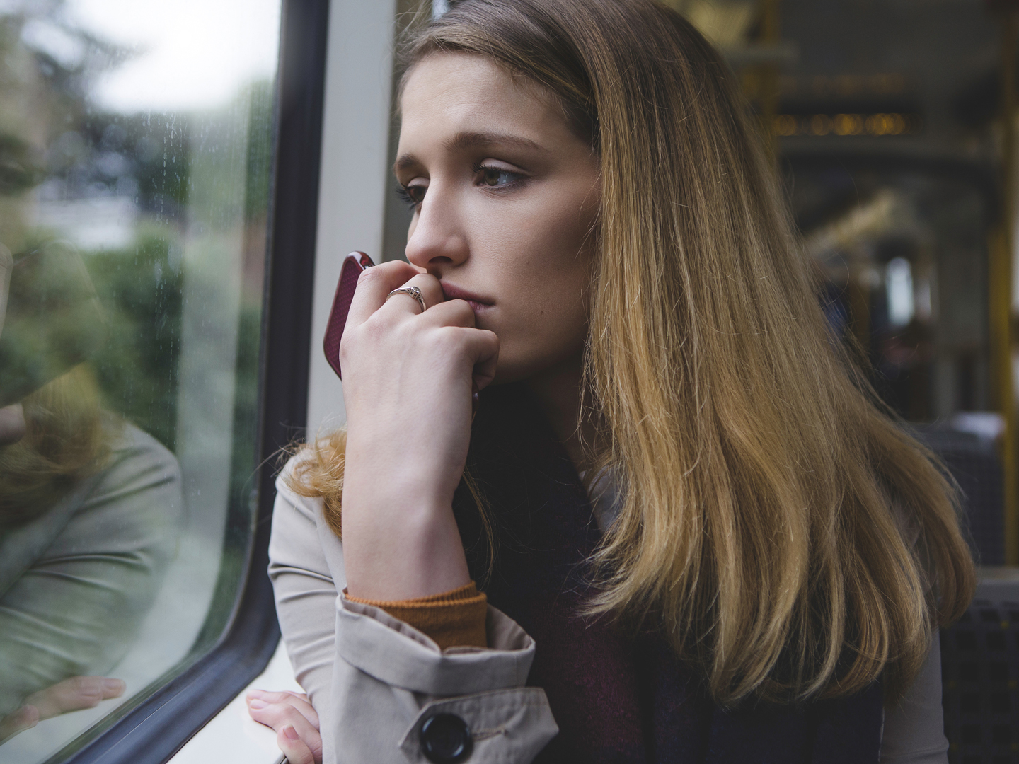 Young woman looking very upset as she sits on a train, looking out of the window. She is holding her phone in her hand up to her mouth.