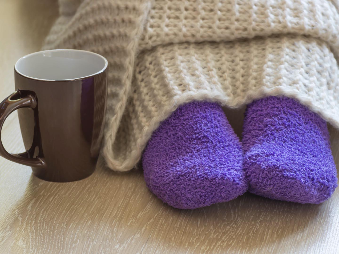 Cold concept with teacup near the legs which are covered with fluffy warm blanket and wearing fluffy warm purple socks