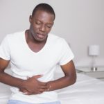 Wincing man sitting on the edge of his bed with a stomach cramp at home in bedroom