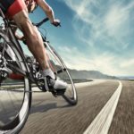 An athlete is riding a bicycle on road. The man is wearing black bike shorts and shin guards along with a red sleeveless top and a red and white helmet and sunglasses. The image is blurred in motion.