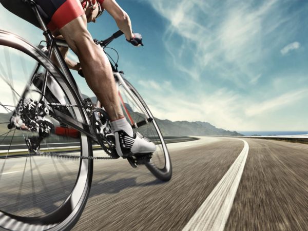 An athlete is riding a bicycle on road. The man is wearing black bike shorts and shin guards along with a red sleeveless top and a red and white helmet and sunglasses. The image is blurred in motion.