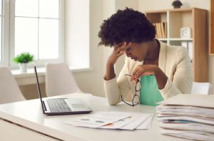Black woman at work with laptop and a stack of files and papers. She has taken off her glasses and is rubbing her head. Overworked.
